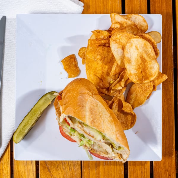 A chicken sandwich with lettuce, tomato, and melted cheese, served with potato chips and a pickle.