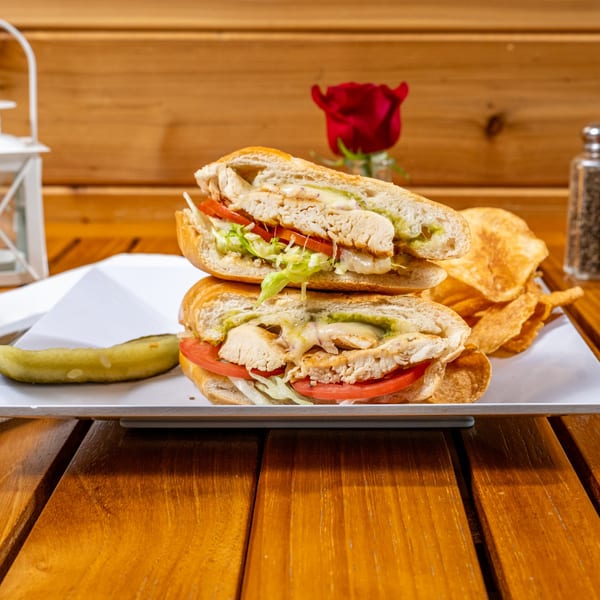 Grilled chicken sandwich with lettuce, tomato, and avocado spread, served with chips and a pickle.