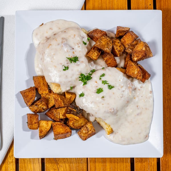 Biscuits and gravy with a side of crispy home fries, garnished with fresh parsley on a white plate.